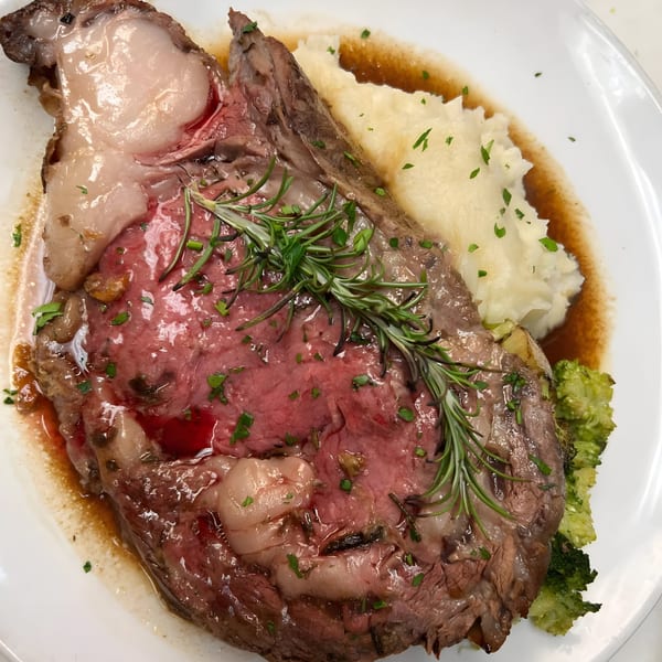 Prime rib with mashed potatoes, gravy, broccoli, and rosemary on a white plate.