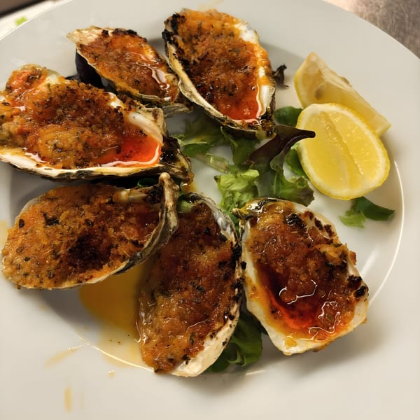 Six baked oysters with breadcrumbs and sauce, garnished with lemon wedges and lettuce on a white plate.