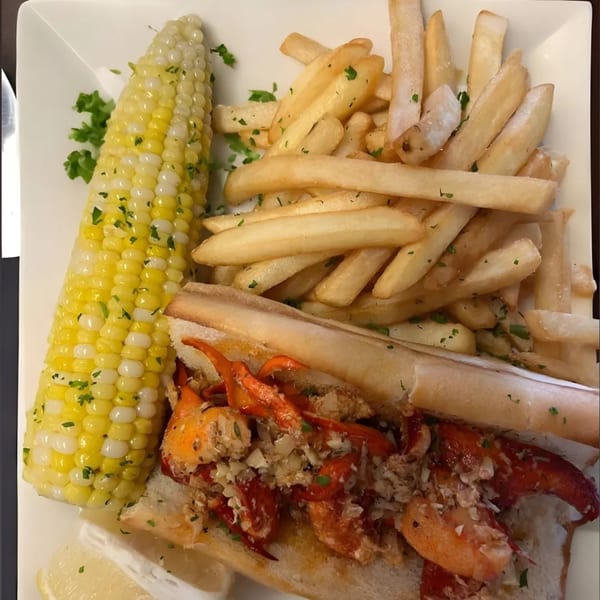 A lobster roll with fries and corn on the cob, garnished with parsley and a lemon wedge.